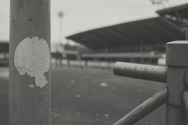 Zdjęcie: stalowy słup latarni przy wejściu na trybunę z wyblakłą naklejką bez napisów, obok zardzewiałe ramię bramki obrotowej. (obraz wygenerowany przez AI)