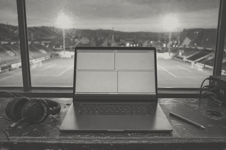 Zdjęcie: laptop z pustymi szarymi wykresami na biurku w loży prasowej, obok ołówek i słuchawki, za oknem pusta murawa o zmierzchu. (obraz wygenerowany przez AI)