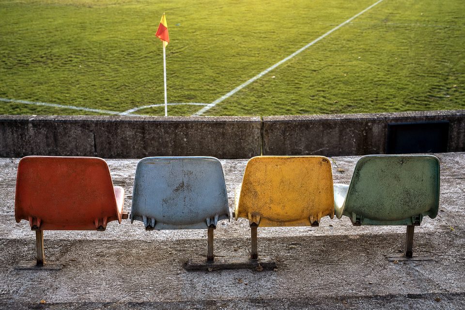 Zdjęcie: cztery zniszczone krzesełka stadionowe w rzędzie, każde w innym wyblakłym kolorze, w tle róg boiska i chorągiewka narożna. (obraz wygenerowany przez AI)
