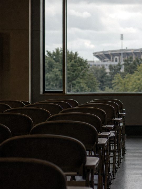 Zdjęcie: rzędy pustych składanych krzeseł w sali wykładowej, w oknie widać dach stadionu i reflektory za drzewami. (obraz wygenerowany przez AI)