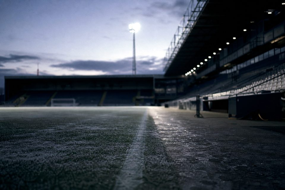 Zdjęcie: zupełnie pusty stadion od linii bocznej w zimowy zmierzch, szron na murawie, krzesełka w cieniu, w oddali świecą reflektory. (obraz wygenerowany przez AI)