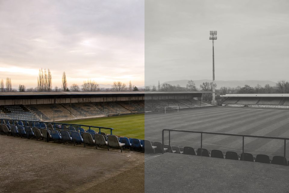 Zdjęcie: pusty stadion niższej ligi o świcie, rzędy zniszczonych krzesełek, murawa w pasy po koszeniu i maszty oświetleniowe. (obraz wygenerowany przez AI)
