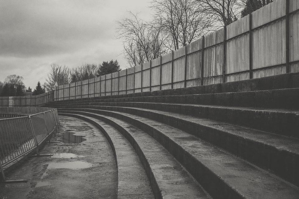 Zdjęcie: zniszczone betonowe stopnie trybuny z wygiętą barierką i wysłużonym metalowym ogrodzeniem, pusto, po deszczu. (obraz wygenerowany przez AI)