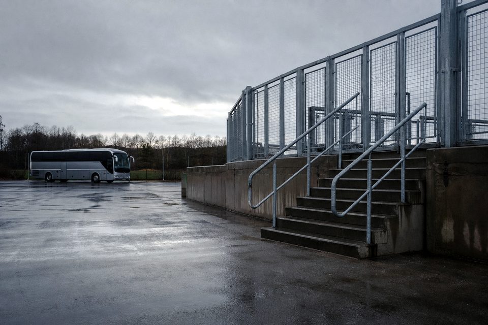 Zdjęcie: klatkowe ogrodzenie sektora gości i puste stalowe schody prowadzące do niego, na skraju parkingu przy stadionie stoi autokar. (obraz wygenerowany przez AI)