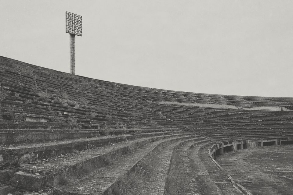 Zdjęcie: rozpadająca się betonowa niecka stadionu z połowy XX wieku, chwasty między stopniami trybun i zniszczony maszt oświetleniowy. (obraz wygenerowany przez AI)