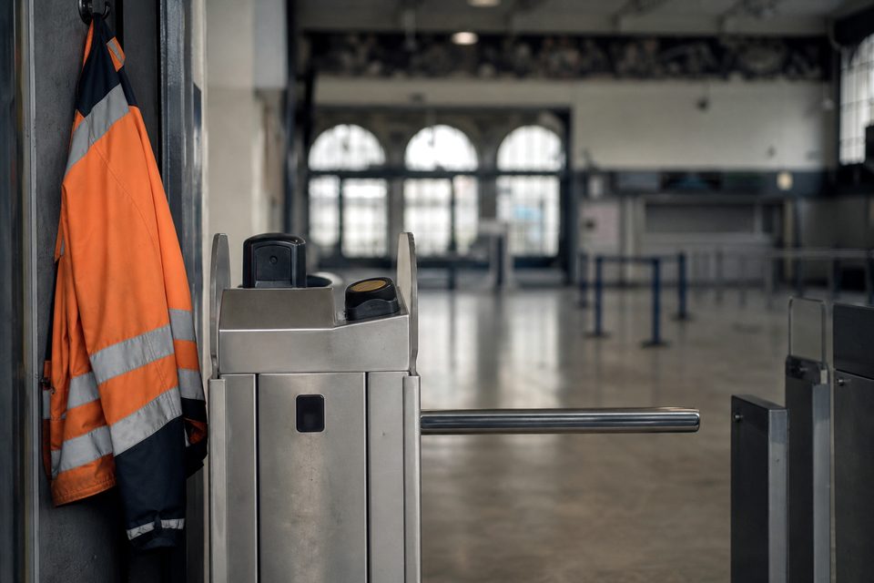 Zdjęcie: zamknięta bramka obrotowa z zablokowanym ramieniem, obok na haku kamizelka odblaskowa stewarda, za nią pusty hall. (obraz wygenerowany przez AI)