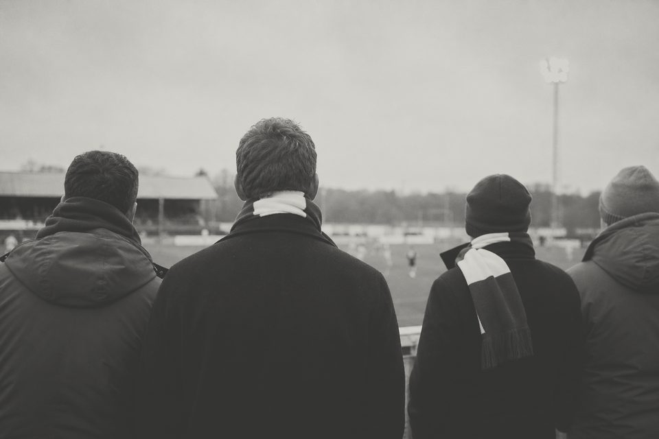 Zdjęcie: kilku kibiców w szalikach widzianych z tyłu z oddali, stojących na trybunie i patrzących na boisko pod szarym niebem. (obraz wygenerowany przez AI)
