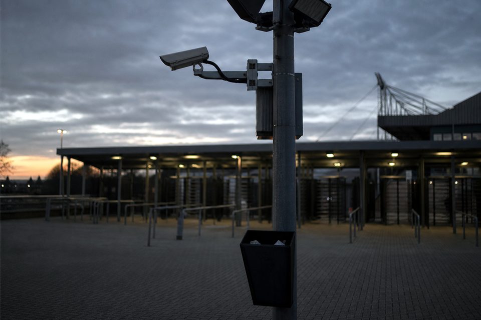 Zdjęcie: uchwyt kamery monitoringu na maszcie oświetleniowym nad wejściem z bramkami obrotowymi, poniżej kosz na papierowe odcinki biletów. (obraz wygenerowany przez AI)