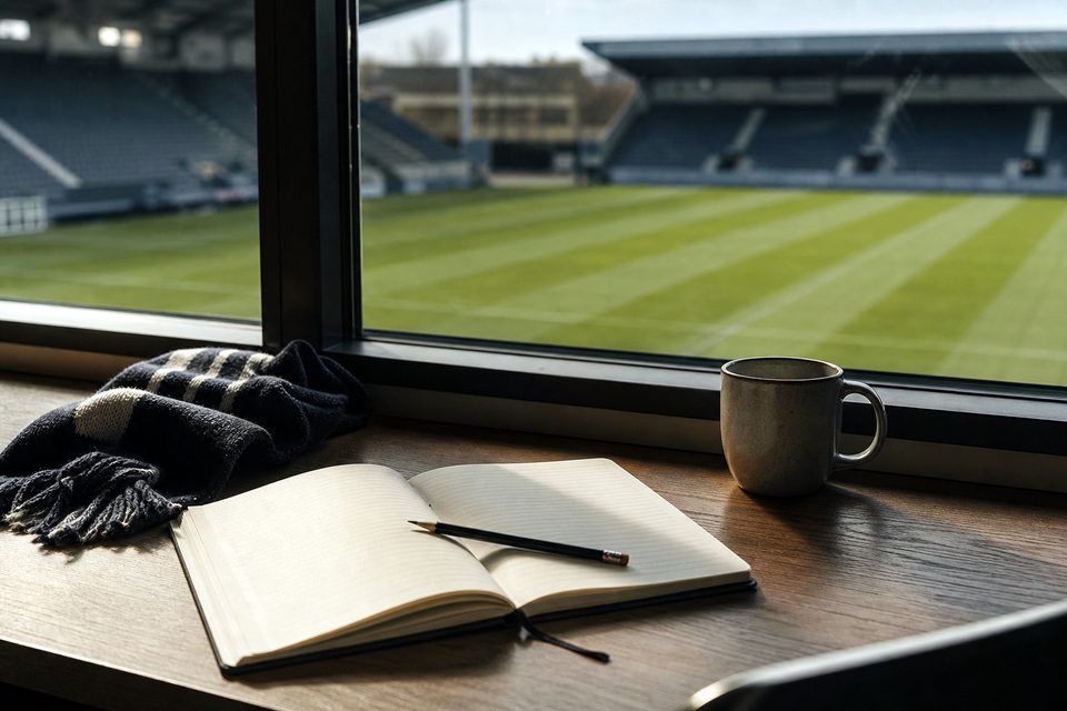 Zdjęcie: redakcyjne biurko przy oknie stadionu z notesem, ołówkiem, kubkiem i szalikiem, za nim nieostra pusta murawa z pasem po kosiarce. (obraz wygenerowany przez AI)