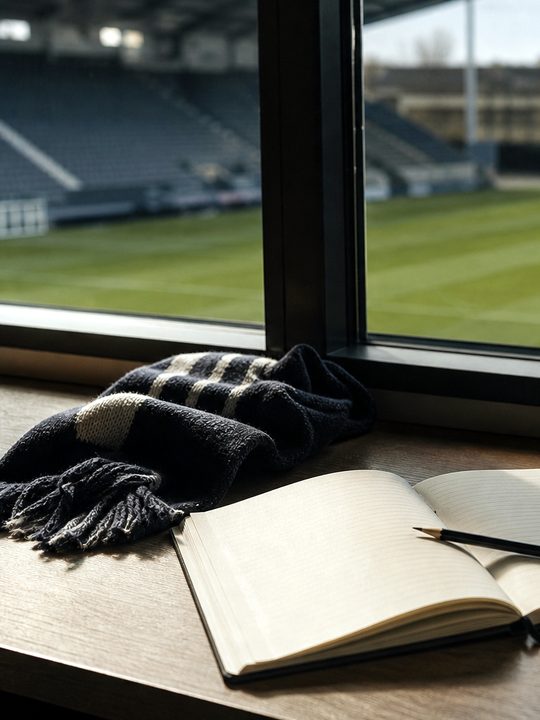 Zdjęcie: redakcyjne biurko przy oknie stadionu z notesem, ołówkiem, kubkiem i szalikiem, za nim nieostra pusta murawa z pasem po kosiarce. (obraz wygenerowany przez AI)