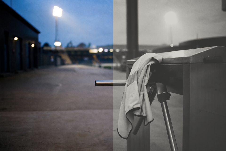 Zdjęcie: stadionowa bramka obrotowa z kamizelką odblaskową stewarda na ramieniu, za nią pusty hall i zapalające się reflektory. (obraz wygenerowany przez AI)