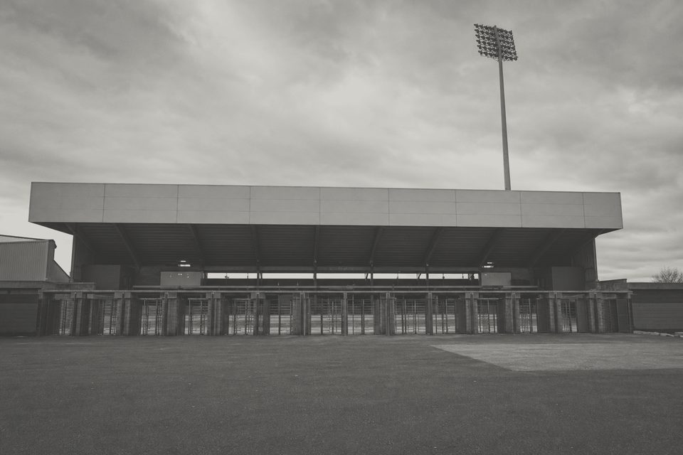 Zdjęcie: pusty przód dachu stadionu z nieoświetlonymi, nieoznakowanymi panelami, maszt oświetleniowy i pusty plac przy bramkach poniżej. (obraz wygenerowany przez AI)