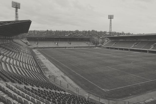 Zdjęcie: panorama średniego stadionu środkowoeuropejskiego z dalekiego rogu, cztery trybuny z różnych epok, maszty oświetleniowe i murawa. (obraz wygenerowany przez AI)