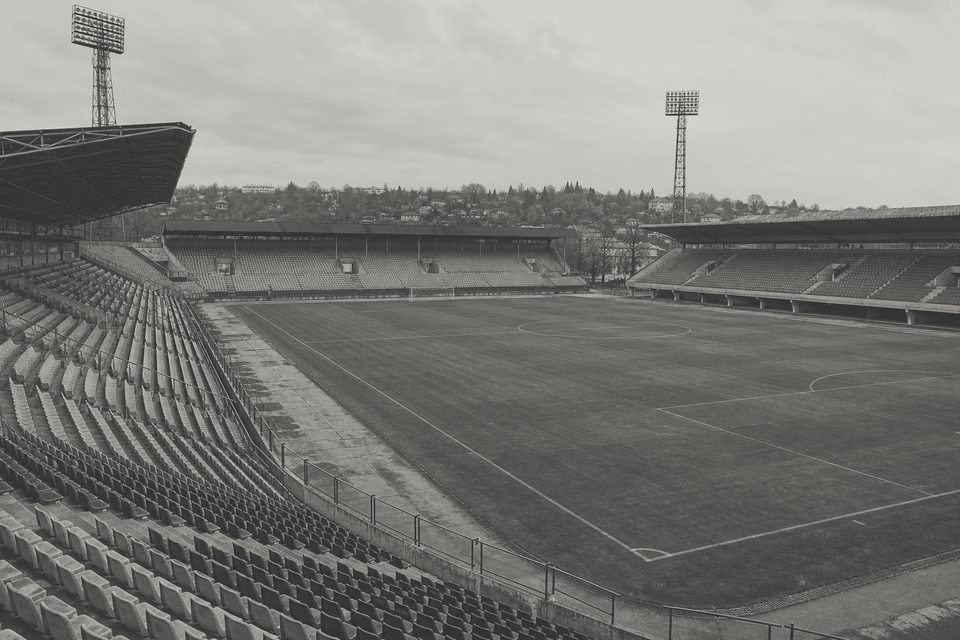 Zdjęcie: panorama średniego stadionu środkowoeuropejskiego z dalekiego rogu, cztery trybuny z różnych epok, maszty oświetleniowe i murawa. (obraz wygenerowany przez AI)