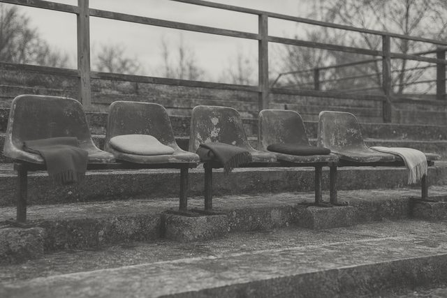Zdjęcie: pięć zniszczonych krzesełek stadionowych w rozstawionej linii na trybunie, przy każdym inny zwykły szalik lub poduszka, bez ludzi. (obraz wygenerowany przez AI)