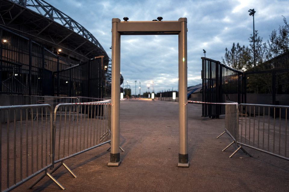Zdjęcie: wejście na stadion z bramką do wykrywania metalu, taśmą zaporową i stalowymi barierkami w perspektywie, pusto, wczesny wieczór. (obraz wygenerowany przez AI)