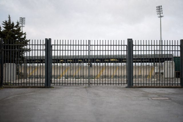 Zdjęcie: wysokie stalowe ogrodzenie i brama widziane sprzed stadionu, przez pręty widać trybunę i reflektory, przed nimi pusty chodnik. (obraz wygenerowany przez AI)